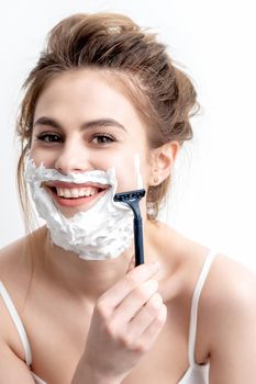 Beautiful young caucasian woman shaving her face by razor on white background. Pretty woman with shaving foam on her face