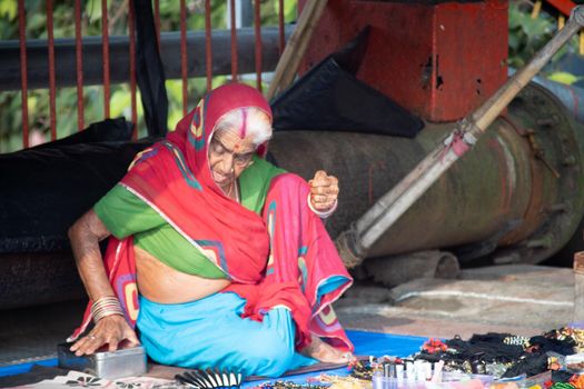 Hadiwar, Uttarakhand, India - circa 2021:Old streetside shopkeeper making handmade bead necklaces for sale sitting under a plastic sheet, haridwar houses a lot of these old destitute people working to make ends meet