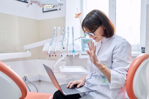 Online consultation, doctor help in clinic using laptop. Female dentist talking waving hand talking looking in laptop in dental office. Video conference, videocall, service, dentistry medicine concept