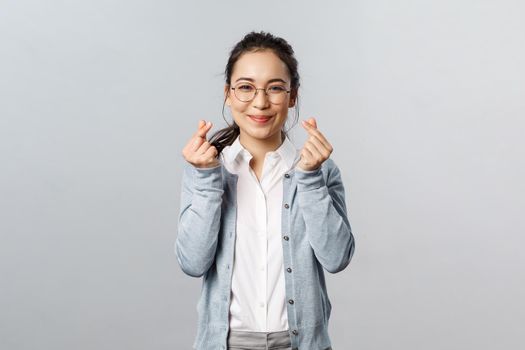 Emotions, people and lifestyle concept. Optimistic, lovely and cute asian girl in glasses express sympathy or joy, showing korean heart signs with fingers, looking kawaii, standing grey background.
