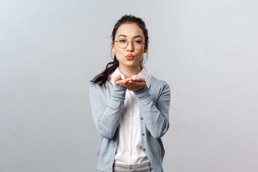 Portrait of romantic and passionate, pretty asian girlfriend in glasses send, blowing air kiss to camera with hands near folded lips, express symapthy, romance and flirt, stand grey background.