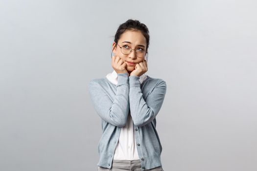 Emotions, people and lifestyle concept. Adorable dreamy and romantic asian girl in glasses, sighing daydreaming, being in love, thinking about dating guy she likes, standing grey background.