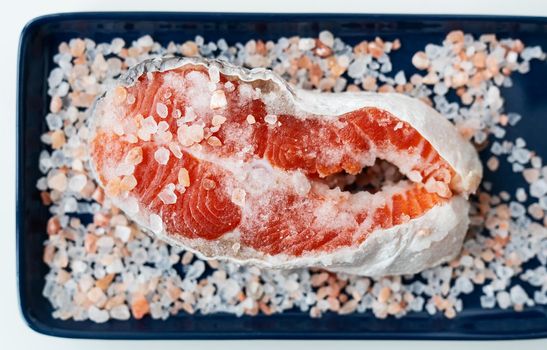 Fresh raw red salmon steak with large coarse pink salt prepared for baking on the grill lies on a blue plate. Healthy seafood food. Top view, place for an inscription