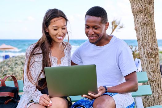 Young couple looking at laptop screen, sitting on bench on the beach, sea nature background. Lifestyle, vacation, summer, family, leisure, relationship, people concept