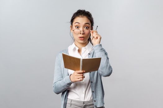 Education, teachers, university and schools concept. Surprised silly asian female student studying, holding notebooks and pen, fixing glasses look wondered at camera, hear question from student.