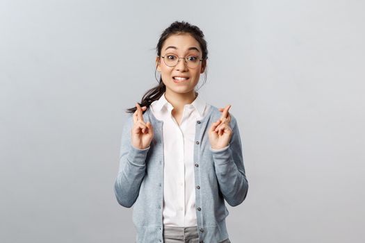 Emotions, people and lifestyle concept. Excited asian girl, biting lip tempting see something, look with aspiration and interest, cross fingers for good luck, praying hopeful over grey background.