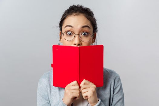 Education, university and people concept. Close-up portrait of surprised and amused young asian girl, widen eyes at amazing interesting story, cover mouth behind red planner, stand grey background.