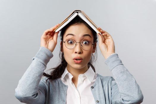 Education, university and people concept. Close-up portrait of wondred and amused asian female in glasses, holding planner, notebook or book on head, realise something interesting, grey background.