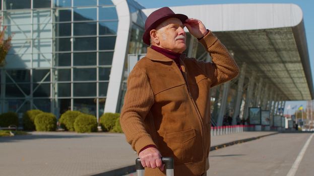 Portrait of elderly retired man tourist near airport terminal waiting boarding on plane for traveling. Stylish senior mature grandfather carrying luggage suitcases bag to railway station. Vacation
