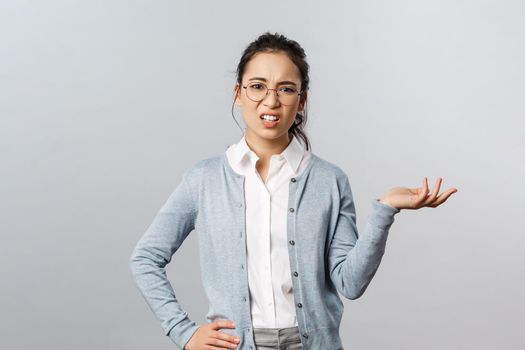 So what. Skeptical arrogant asian woman, looking with dismay and judgement camera, raise hand sideways and shrugging unbothered, look frustrated at something awful and bad, grey background.