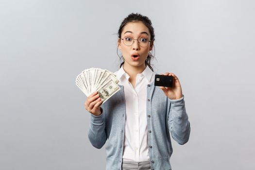Business, finance and investment concept. Amazed curious asian female in glasses, look impressed and surprised, holding dollars cash and credit card, decide how better keep money safe.