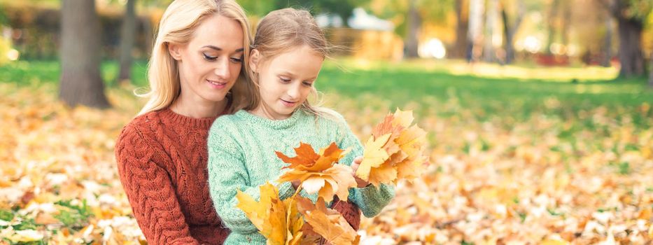 Happy young caucasian woman and little girl holding autumn yellow leaves sitting at the park