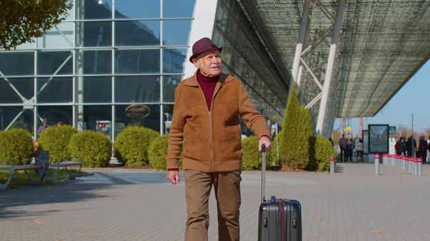 Senior pensioner tourist grandfather businessman walking away from international airport hall or railway station while carrying luggage suitcase on wheels. Travel, vacation, journey, trip, holidays