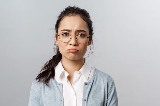 Close-up portrait of gloomy, upset and disappointed young asian female facing failure, losing, regret mjssing interesting event, pouting and sob, frowning camera standing grey background.