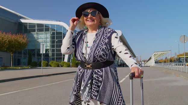 Lovely stylish mature retired grandmother businesswoman walking with luggage suitcase bag to airport hall or railway station for boarding. Travel, vacation, journey, trip. Senior pensioner woman