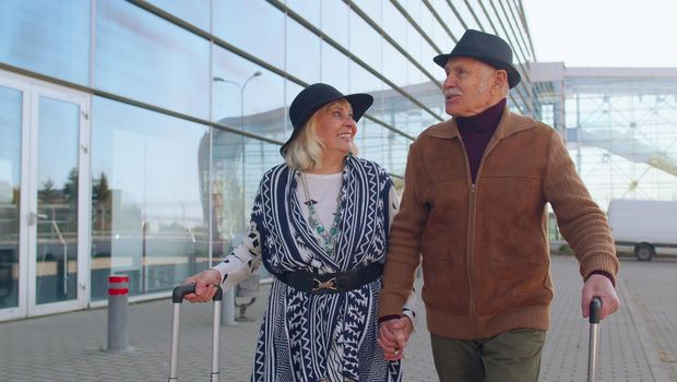 Elderly old husband and wife retirees tourists go to airport terminal for boarding or railway station for traveling. Lovely mature couple grandmother grandfather carrying luggage suitcases on wheels