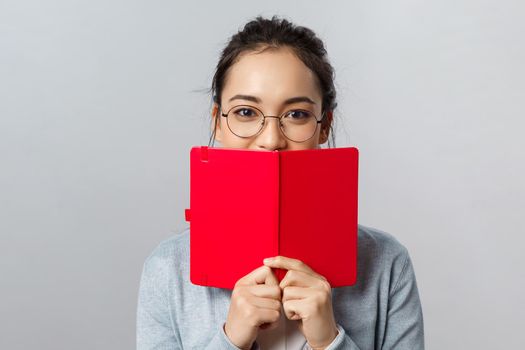 Education, university and people concept. Close-up portrait of romantic young female student, asian girl have secret diary, hiding smile behind planner or notebook, writing notes, grey background.