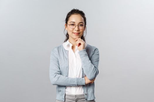 Hmm very interesting. Portrait of intrigued cute asian woman making choice, deciding between variants, look camera satisfied and smile, made-up plan, listening to curious suggestion, grey background.