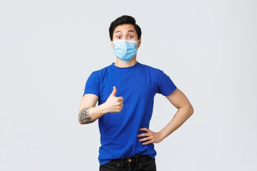 Different emotions, social distancing, self-quarantine on covid-19 and lifestyle concept. Surprised and impressed supportive asian guy thumbs-up to good idea, wearing medical mask.