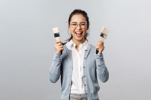 Creativity, repairs and overhaul concept. Excited happy, smiling asian woman holding two painting brushes and laughing with rejoice, cant wait to pain her room new color, have overhaul.