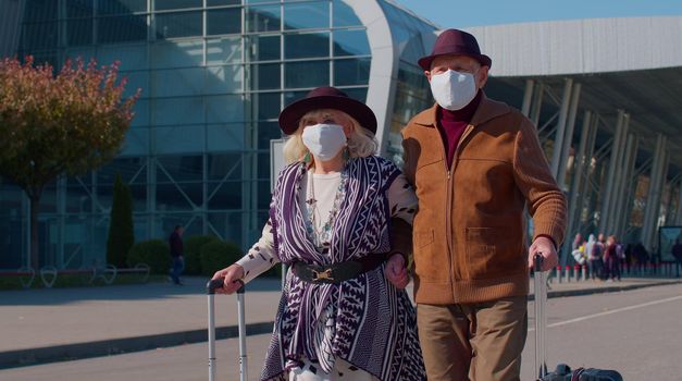 Elderly couple pensioner tourists grandmother grandfather walking with luggage from airport hall or railway station wearing Protective Face Mask COVID-19 coronavirus infection, pandemic virus disease