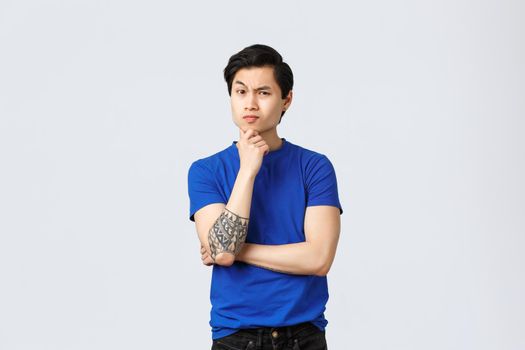Skeptical, serious-looking asian man in blue t-shirt with tattoos, listening suspicious to person, frowning, touch chin thoughtful, thinking, making decision, express disbelief, grey background.