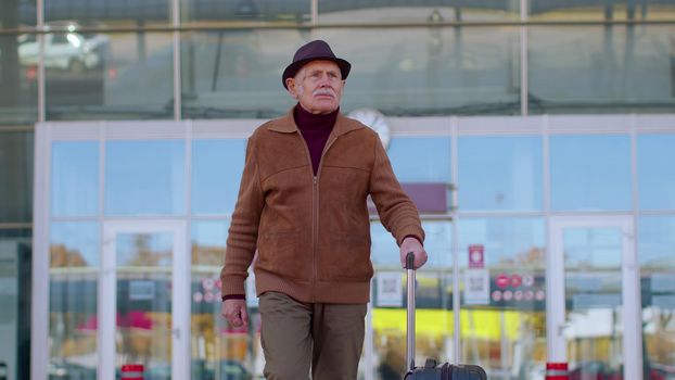 Senior pensioner tourist grandfather businessman walking away from international airport hall or railway station while carrying luggage suitcase on wheels. Travel, vacation, journey, trip, holidays