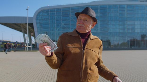 Senior rich pensioner tourist grandfather businessman stay near international airport hall or railway station holding cash money in dollar banknotes celebrate success win. Travel, vacation, holidays