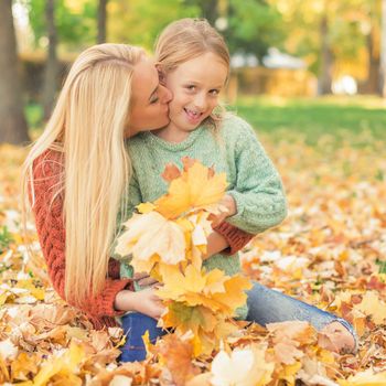 Happy young caucasian mother and little daughter holding autumn yellow leaves sitting and kissing at the park