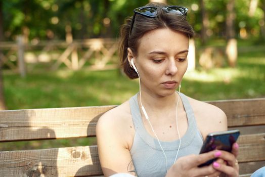 Cheerful millennial blonde teenage girl, sitting on bench outdoors, with smart phone and headphones. Young female student in park smiling and relaxing.