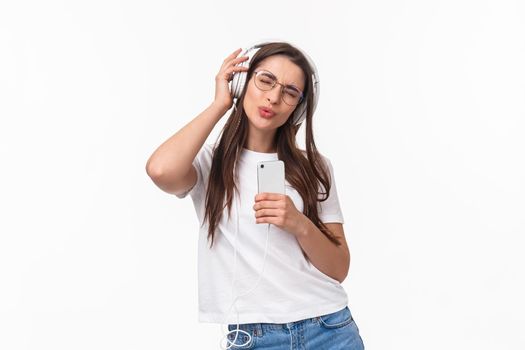 Technology, lifestyle and music concept. Portrait of upbeat, enthusiastic young girl in glasses, listening songs in headphones, singing along awesome track, hold mobile phone and dancing.