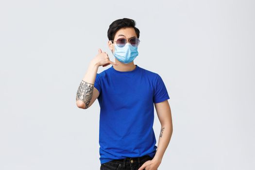 Covid019 lifestyle, people emotions and leisure on quarantine concept. Handsome self-assured young asian guy, macho man in sunglasses and medical mask, flirting, make phone call sign.