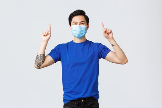 Different emotions, social distancing, self-quarantine on coronavirus and lifestyle concept. Cheerful smiling asian man in medical mask and t-shirt, pointing fingers up to advertise, showing banner.