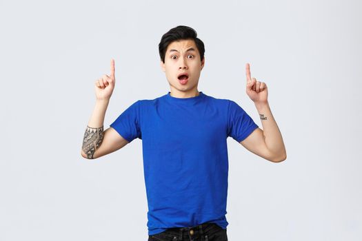 Different emotions, people lifestyle and advertising concept. Surprised and astonished asian male student, guy in blue t-shirt gasping amazed, pointing fingers up at banner, grey background.