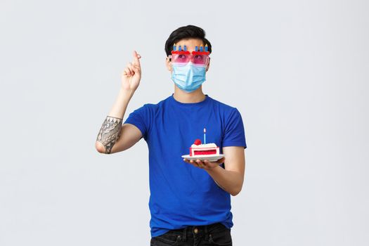 Different emotions, social distancing, self-quarantine on covid-19 and lifestyle concept. Young cute asian guy in funny glasses, medical mask, holding birthday cake and cross fingers making wish.