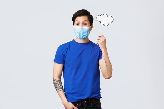 Lifestyle, people different emotions and covid-19 pandemic concept. Surprised young asian man have idea, wear medical mask and hold comment cloud near head, think-up solution.