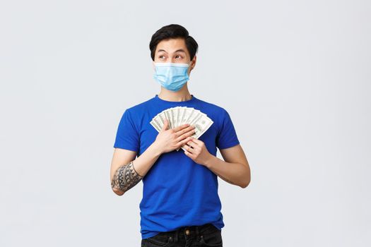 Money, lifestyle, insurance and investment concept. Dreamy happy asian man holding cash and thinking what buy on it, looking away smiling, wear medical mask, grey background.