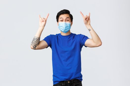 Different emotions, social distancing, self-quarantine on covid-19 and lifestyle concept. Cheerful guy rocking his isolation at home with awesome music, show heavy metal sign, wear medical mask.