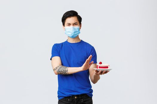 Self-quarantine, home lifestyle and celebration concept. Asian disappointed man celebrating birthday, rejecting piece cake not in mood, being angry standing in medical mask grey background.