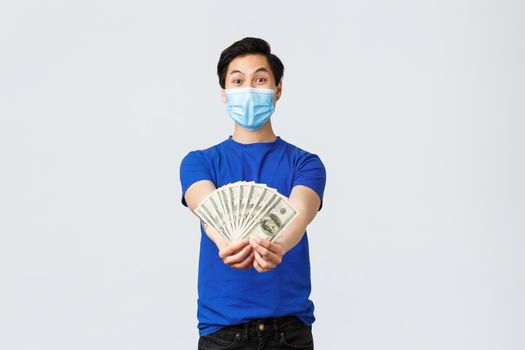 Money, lifestyle, insurance and investment concept. Look how much cash I got. Asian excited guy in medical mask show-off or bragging, showing dollars to you, standing grey background.