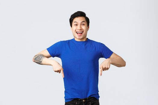 Different emotions, people lifestyle and advertising concept. Enthusiastic handsome asian man in blue t-shirt, pointing fingers down and smiling as announce good news, grey background.