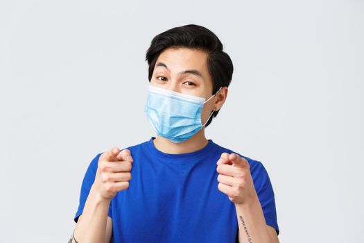 Different emotions, lifestyle and leisure during coronavirus, covid-19 concept. Close-up of attractive asian male in medical mask, guy smiling and pointing at camera, choosing.
