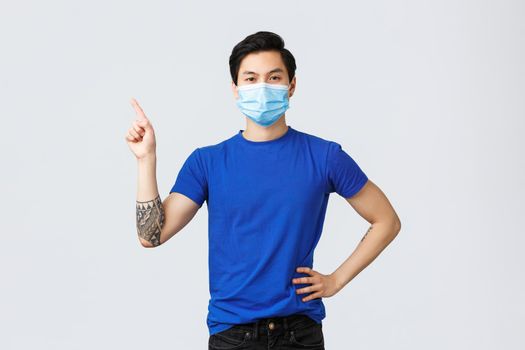 Different emotions, social distancing, self-quarantine on coronavirus and lifestyle concept. Confident asian guy in medical mask pointing finger upper left corner, provide information on covid-19.