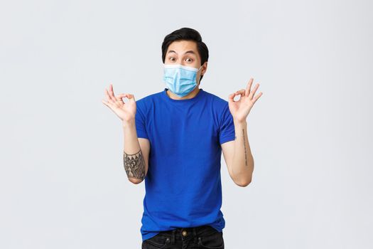 Different emotions, social distancing, self-quarantine on covid-19 and lifestyle concept. Amused happy asian guy in medical mask, show okay sign and smiling, great idea, awesome plan.