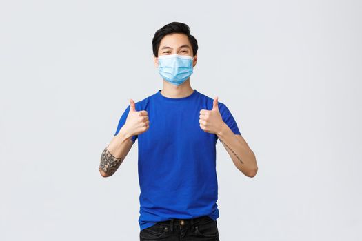 Different emotions, social distancing, self-quarantine on covid-19 and lifestyle concept. Cheerful and supportive young asian guy in medical mask encourage use personal protective equipment.