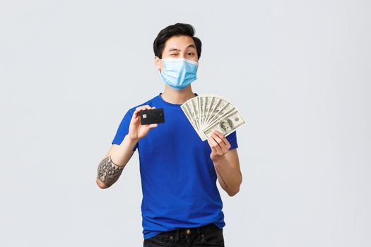 Money, covid-19, easy payment, investment and banking concept. Handsome asian young man assure you need this, showing credit card and cash dollars, wink camera hinting, wear medical mask.