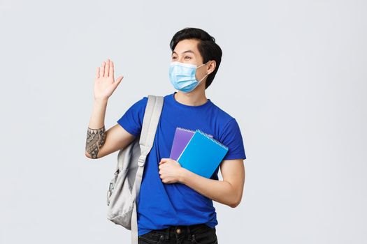 Back to school, studying during covid-19, education and university life concept. Carefree smiling male student waving hand at friend college, head campus or lecture, wear backpack and medical mask.