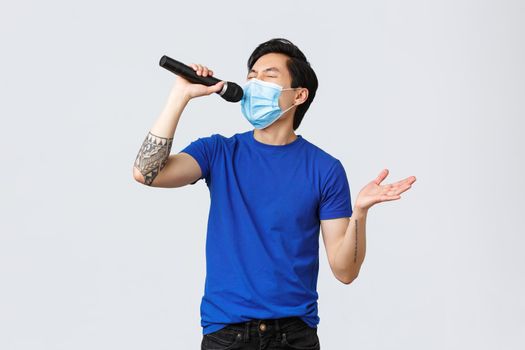 Covid-19 lifestyle, people emotions and leisure on quarantine concept. Handsome funny young male student staying home during coronavirus pandemic, wear mask and singing karaoke in microphone.
