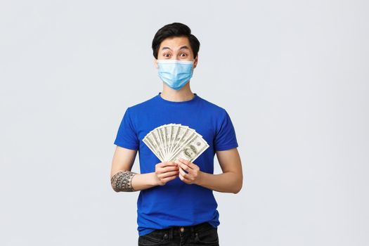 Money, lifestyle, insurance and investment concept. Surprised excited asian man won lottery, earned lots cash, showing dollars and smiling amazed, wearing medical mask.