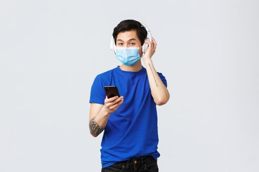 Covid-19 lifestyle, people emotions and leisure on quarantine concept. Handsome asian hipster in medical mask, using headphones listen music, put song on smartphone, hold mobile phone.
